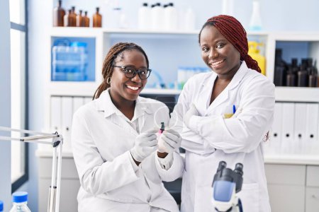 Foto de Mujeres científicas afroamericanas mirando muestra de pie con los brazos cruzados gesto en el laboratorio - Imagen libre de derechos