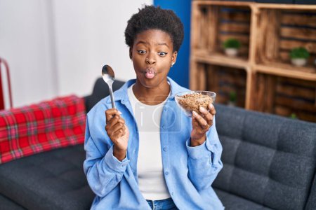 Foto de Mujer afroamericana comiendo cereales integrales saludables haciendo cara de pescado con boca y ojos entrecerrados, locos y cómicos. - Imagen libre de derechos