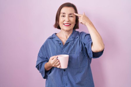 Foto de Mujer hispana de mediana edad bebiendo una taza de café sonriendo señalando a la cabeza con un dedo, gran idea o pensamiento, buena memoria - Imagen libre de derechos