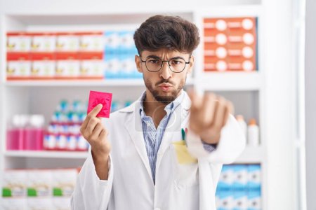 Téléchargez les photos : Homme arabe avec barbe travaillant à la pharmacie pharmacie tenant préservatif faisant visage de poisson avec la bouche et les yeux plissés, fou et comique. - en image libre de droit
