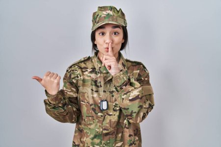 Foto de Joven mujer del sur de Asia con camuflaje uniforme del ejército pidiendo estar tranquilo con el dedo en los labios señalando con la mano a un lado. silencio y concepto secreto. - Imagen libre de derechos
