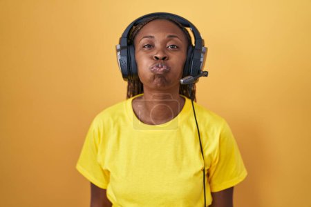 Téléchargez les photos : Femme afro-américaine écoutant de la musique à l'aide d'écouteurs gonflant les joues avec un visage drôle. bouche gonflée d'air, expression folle. - en image libre de droit