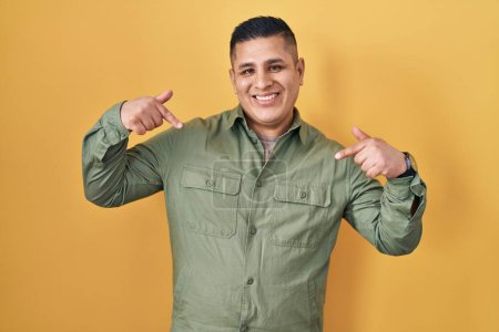 Foto de Joven hispano parado sobre fondo amarillo mirando confiado con sonrisa en la cara, señalándose con los dedos orgullosos y felices. - Imagen libre de derechos