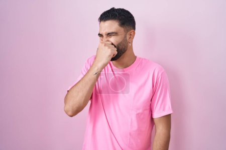 Téléchargez les photos : Jeune homme hispanique debout sur fond rose sentant quelque chose de puant et dégoûtant, odeur intolérable, retenant souffle avec les doigts sur le nez. mauvaise odeur - en image libre de droit