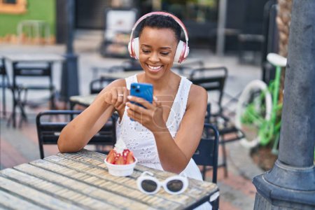 Photo for African american woman eating ice cream listening to music at coffee shop terrace - Royalty Free Image