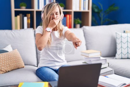 Téléchargez les photos : Jeune femme blonde étudiant à l'aide d'un ordinateur portable à la maison se moquant de vous, pointant du doigt la caméra avec la main sur la bouche, expression de honte - en image libre de droit