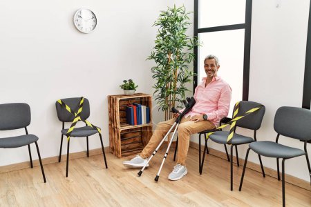 Téléchargez les photos : Homme aux cheveux gris d'âge moyen blessé assis sur une chaise tenant des béquilles dans la salle d'attente de la clinique - en image libre de droit