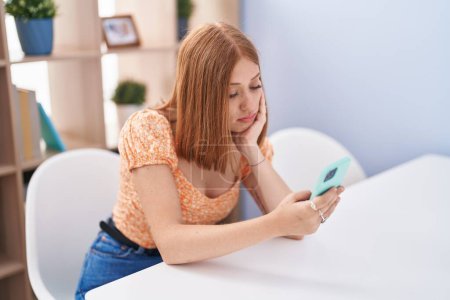 Téléchargez les photos : Jeune rousse femme en utilisant smartphone assis sur la table à la maison - en image libre de droit