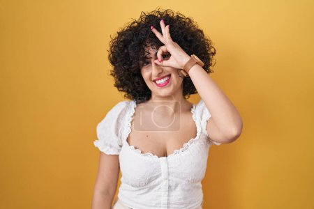Photo for Young brunette woman with curly hair standing over yellow background doing ok gesture with hand smiling, eye looking through fingers with happy face. - Royalty Free Image