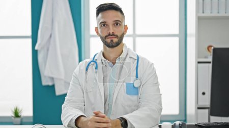 Photo for Young hispanic man doctor sitting on table with serious expression at the clinic - Royalty Free Image