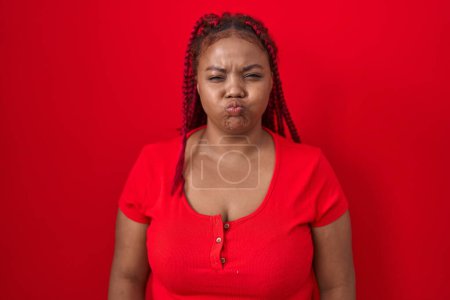 Foto de Mujer afroamericana con el pelo trenzado de pie sobre fondo rojo hinchando mejillas con cara divertida. boca hinchada de aire, expresión loca. - Imagen libre de derechos