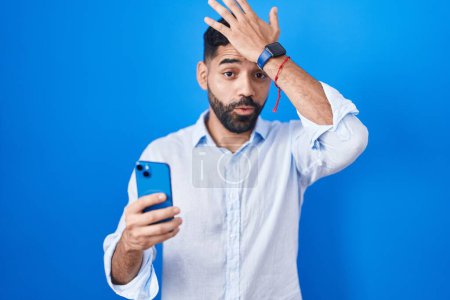 Téléchargez les photos : Homme hispanique avec barbe en utilisant le message de dactylographie smartphone surpris avec la main sur la tête pour erreur, rappelez-vous erreur. oublié, mauvais concept de mémoire. - en image libre de droit