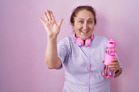 Foto de Mujer mayor con ropa deportiva y auriculares renunciando a decir hola feliz y sonriente, gesto de bienvenida amistoso - Imagen libre de derechos