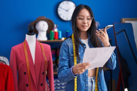 Téléchargez les photos : Jeune femme chinoise tailleur parler sur smartphone document de lecture à l'usine de vêtements - en image libre de droit