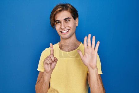 Téléchargez les photos : Jeune homme debout sur fond bleu montrant et pointant vers le haut avec les doigts numéro six tout en souriant confiant et heureux. - en image libre de droit