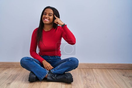 Foto de Joven afroamericano con trenzas sentado en el suelo en casa sonriendo apuntando a la cabeza con un dedo, gran idea o pensamiento, buena memoria - Imagen libre de derechos