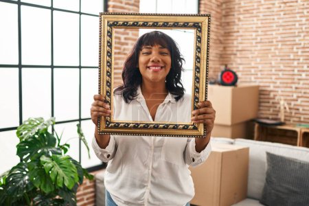 Photo for Hispanic woman at new home holding empty frame smiling with a happy and cool smile on face. showing teeth. - Royalty Free Image