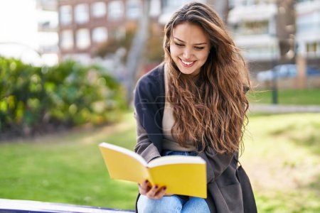 Foto de Joven mujer hispana hermosa leyendo libro sentado en el banco en el parque - Imagen libre de derechos