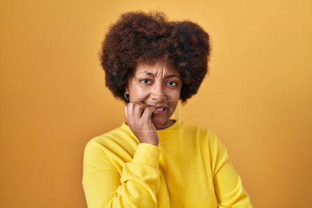 Téléchargez les photos : Jeune femme afro-américaine debout sur fond jaune regardant stressé et nerveux avec les mains sur les ongles mordants de la bouche. problème d'anxiété. - en image libre de droit