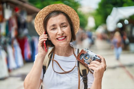 Photo for Middle age woman tourist talking on smartphone holding camera at street market - Royalty Free Image