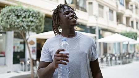 Téléchargez les photos : Un homme afro-américain tenant une bouteille d'eau regardant le ciel souriant sur la terrasse du café - en image libre de droit