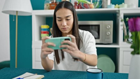 Photo for Young beautiful hispanic woman playing video game by smartphone at dinning room - Royalty Free Image