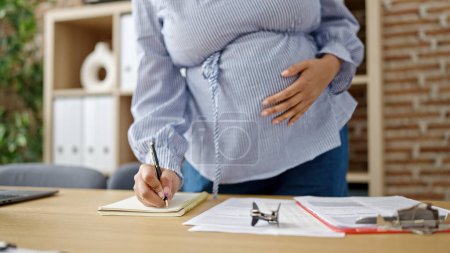 Photo for Young pregnant woman business worker writing on notebook touching belly at office - Royalty Free Image