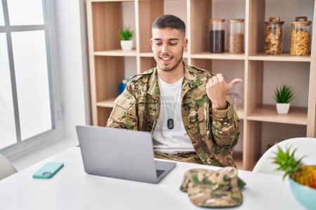 Photo for Young hispanic man wearing camouflage army uniform doing video call pointing thumb up to the side smiling happy with open mouth - Royalty Free Image