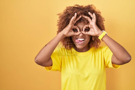 Foto de Mujer hispana joven con el pelo rizado de pie sobre el fondo amarillo haciendo buen gesto como prismáticos sacando la lengua, los ojos mirando a través de los dedos. expresión loca. - Imagen libre de derechos