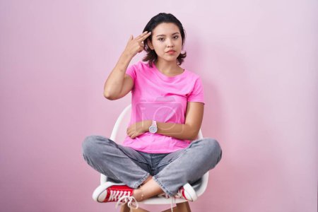 Foto de Mujer joven hispana sentada en silla sobre fondo rosa disparando y suicidándose apuntando de la mano y los dedos a la cabeza como arma, gesto suicida. - Imagen libre de derechos