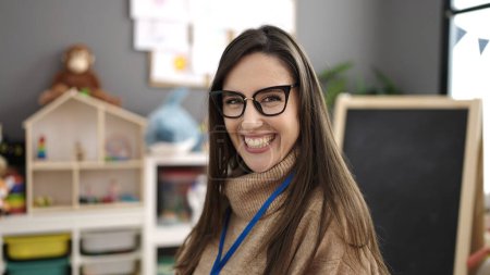 Téléchargez les photos : Belle femme hispanique prof préscolaire souriant confiance debout à la maternelle - en image libre de droit