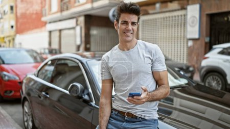 Photo for Young hispanic man using smartphone sitting on car smiling at street - Royalty Free Image