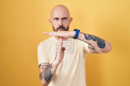 Foto de Hombre hispano con tatuajes de pie sobre fondo amarillo haciendo un gesto de tiempo fuera con las manos, rostro frustrado y serio - Imagen libre de derechos