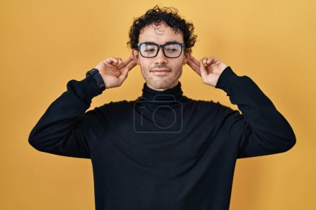 Téléchargez les photos : Homme hispanique debout sur fond jaune souriant tirant les oreilles avec les doigts, geste drôle. problème d'audition - en image libre de droit