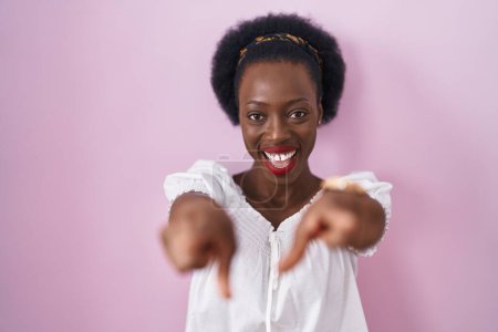 Téléchargez les photos : Femme africaine avec les cheveux bouclés debout sur fond rose pointant vers vous et la caméra avec les doigts, souriant positif et joyeux - en image libre de droit