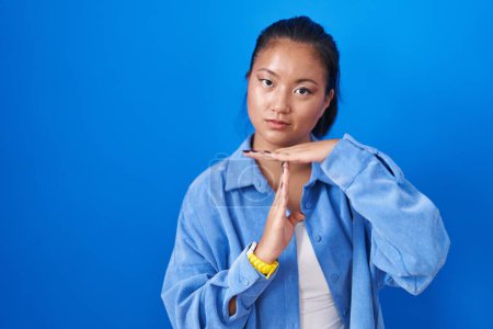 Foto de Asiática joven mujer de pie sobre fondo azul haciendo tiempo fuera gesto con las manos, frustrado y serio rostro - Imagen libre de derechos