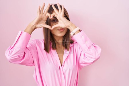 Foto de Mujer hispana joven con gafas de pie sobre fondo rosa haciendo forma de corazón con la mano y los dedos sonriendo mirando a través de signo - Imagen libre de derechos