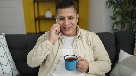 Foto de Un joven hispano hablando por teléfono bebiendo una taza de café en casa - Imagen libre de derechos