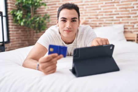 Téléchargez les photos : Jeune homme hispanique utilisant touchpad et carte de crédit assis sur le lit dans la chambre - en image libre de droit