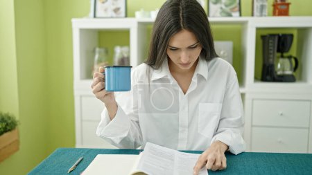 Foto de Joven hermosa mujer hispana leyendo documento bebiendo café en el comedor - Imagen libre de derechos