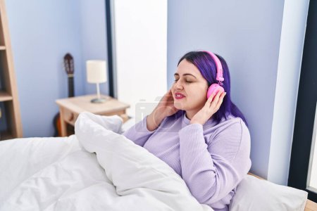 Foto de Joven hermosa mujer de tamaño grande escuchando música sentada en la cama en el dormitorio - Imagen libre de derechos