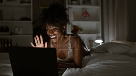 Foto de Mujer afroamericana vistiendo lencería teniendo videollamada acostada en la cama en el dormitorio - Imagen libre de derechos