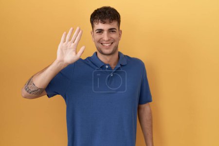 Foto de Joven hombre hispano de pie sobre fondo amarillo renunciando a decir hola feliz y sonriente, gesto de bienvenida amistoso - Imagen libre de derechos