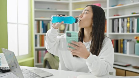 Téléchargez les photos : Jeune belle étudiante hispanique utilisant un smartphone boire une boisson énergétique à la salle de classe de l'université - en image libre de droit