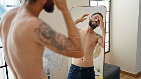 Téléchargez les photos : Jeune homme hispanique peigner les cheveux avec les mains regardant sur le miroir à la maison - en image libre de droit
