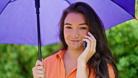 Téléchargez les photos : Jeune belle femme hispanique tenant parapluie parlant sur smartphone au parc - en image libre de droit