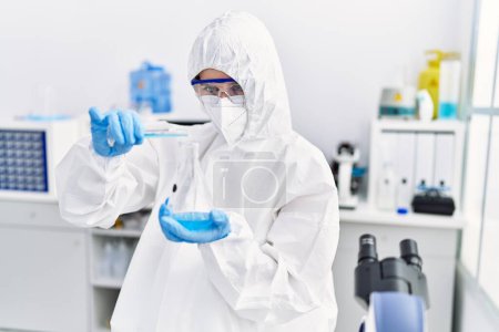 Foto de Joven científica rubia vistiendo uniforme de seguridad vertiendo líquido en el tubo de ensayo en el laboratorio - Imagen libre de derechos