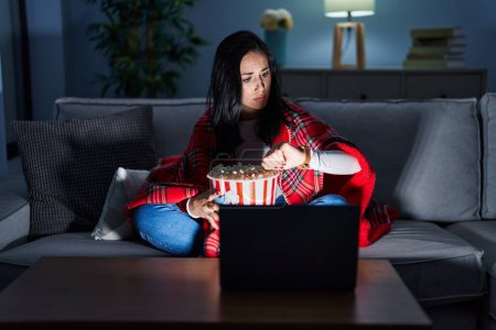 Photo for Hispanic woman eating popcorn watching a movie on the sofa checking the time on wrist watch, relaxed and confident - Royalty Free Image