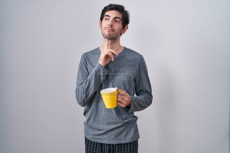 Photo for Young hispanic man wearing pajama drinking a cup of coffee thinking concentrated about doubt with finger on chin and looking up wondering - Royalty Free Image
