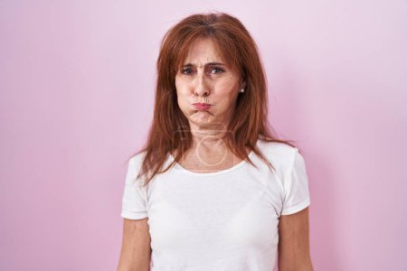 Foto de Mujer de mediana edad de pie sobre fondo rosa hinchando mejillas con cara divertida. boca hinchada de aire, expresión loca. - Imagen libre de derechos